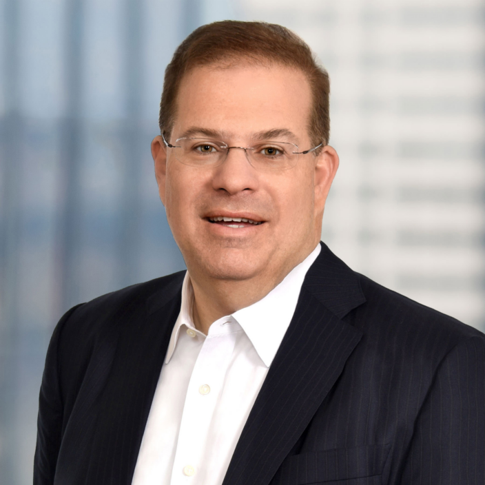 A man wearing glasses, a dark pinstripe suit, and a white dress shirt, smiling at the camera with a blurred office background.