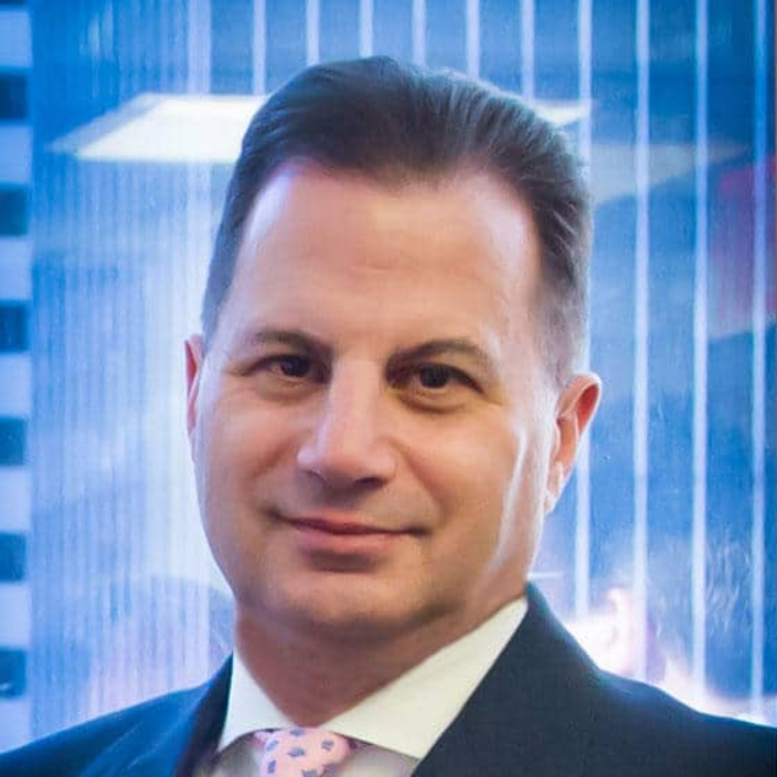 A man in a suit and pink patterned tie stands in front of a window with tall buildings in the background, smiling slightly at the camera.