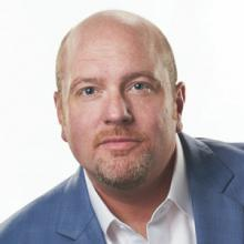 A bald man with light skin, light facial hair, and blue eyes, wearing a light blue suit jacket and white shirt, looks directly at the camera against a plain white background.