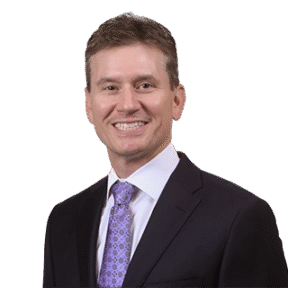 A man in a dark suit, white shirt, and patterned purple tie smiles at the camera against a plain white background.