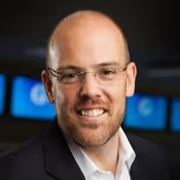 A smiling man with a bald head, glasses, and a trimmed beard, wearing a dark suit jacket and white shirt, with blurred blue screens in the background.