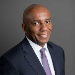 A man in a dark suit, white shirt, and striped tie smiles at the camera against a plain gray background.