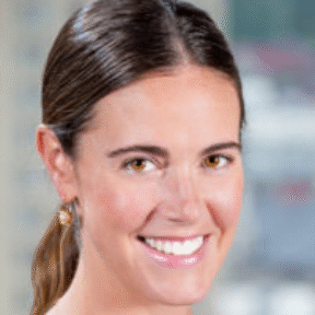 A woman with long brown hair tied back, wearing a sleeveless black top, small gold earrings, and a delicate necklace, smiles at the camera. The background is blurred with buildings visible.