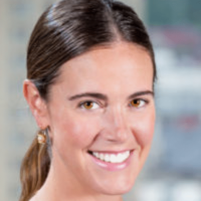 A woman with long brown hair tied back, wearing a sleeveless black top, small gold earrings, and a delicate necklace, smiles at the camera. The background is blurred with buildings visible.