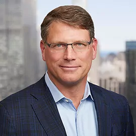 A man with short brown hair and glasses, wearing a blue plaid suit jacket and a light blue shirt, stands in front of a blurred cityscape background.
