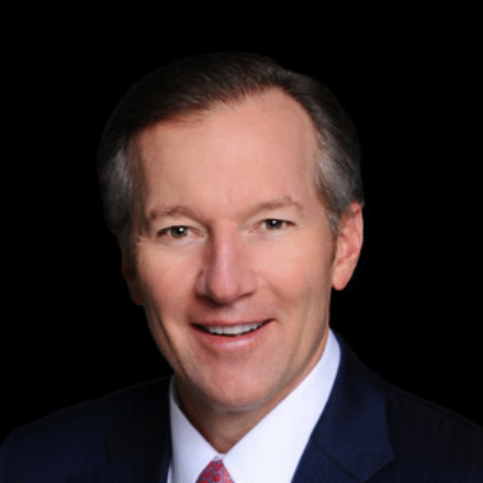 A middle-aged man with short brown hair wearing a dark blue suit, white shirt, and red patterned tie, smiling against a black background.