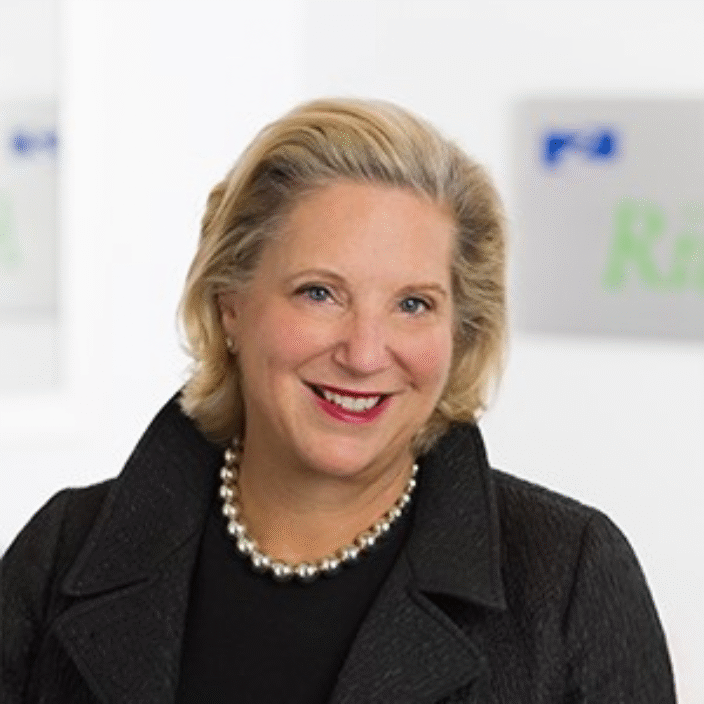 A woman with blonde hair wearing a black textured coat, a black top, and a pearl necklace, smiling in front of a blurred background with partially visible signage.