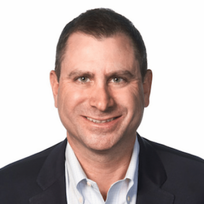 A man with short brown hair, wearing a dark blazer over a light-colored collared shirt, smiles at the camera against a plain white background.