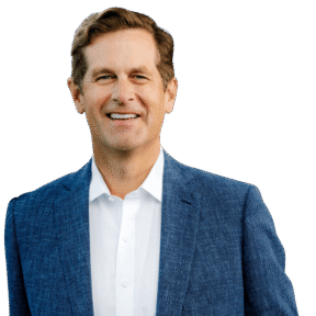 A man with short brown hair wearing a blue textured suit jacket over a white dress shirt stands smiling against a plain white background.