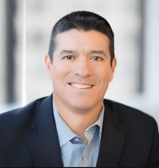 A man with short dark hair wearing a dark blazer and light blue shirt smiles at the camera in a bright, blurred office setting.