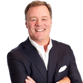 Smiling man with short light brown hair in a dark suit and white dress shirt, arms crossed, standing against a plain light background.