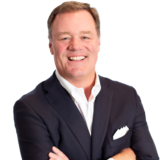 Smiling man with short light brown hair in a dark suit and white dress shirt, arms crossed, standing against a plain light background.