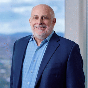 A smiling, bald man with a gray beard wears a blue blazer and light blue checked shirt, standing in front of a window with a blurred outdoor background.