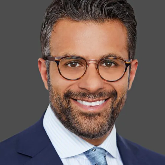 A man with short, dark hair, a beard, and glasses smiles while wearing a navy blue suit, light blue shirt, and patterned tie against a plain grey background.