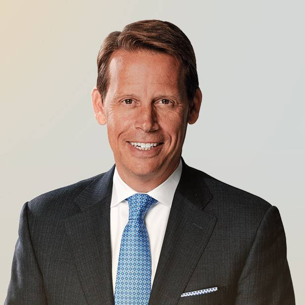 A man in a dark suit, white shirt, and blue patterned tie is smiling at the camera against a plain, light-colored background.