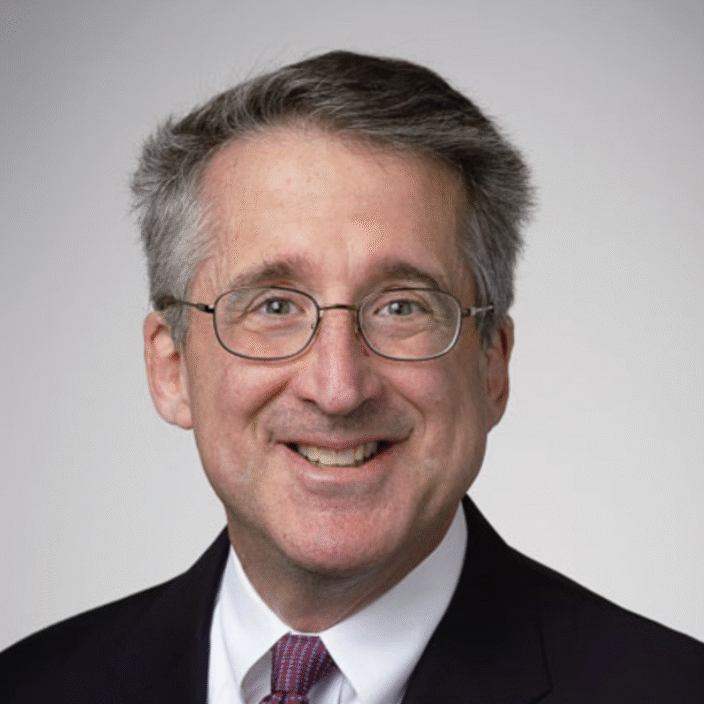 A middle-aged man with gray hair, glasses, and a friendly smile is wearing a dark suit jacket, white dress shirt, and a red patterned tie. The background is plain and light-colored.