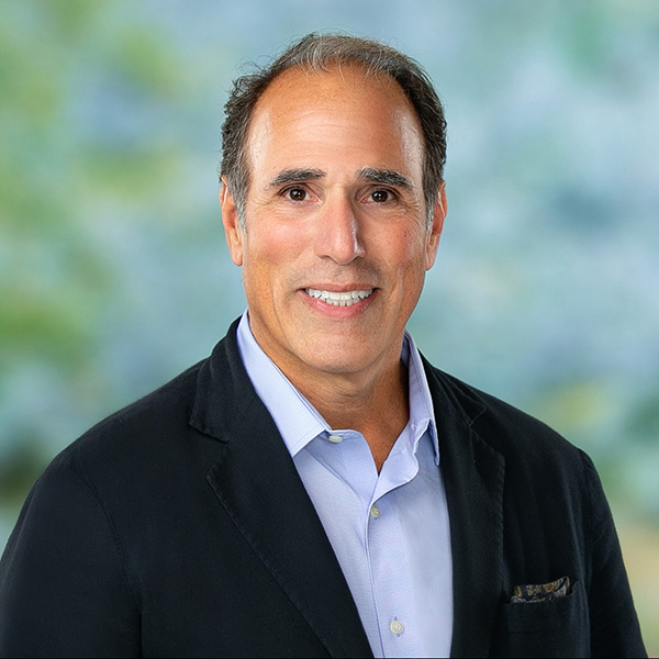 A middle-aged man with short dark hair, wearing a black blazer over a light blue shirt, smiles at the camera against a blurred outdoor background.