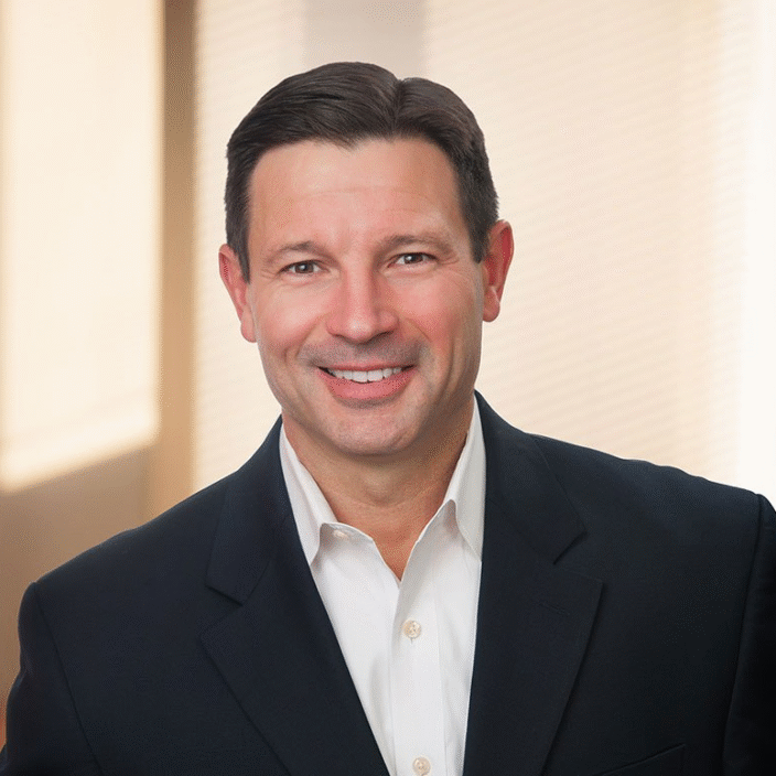A man with short dark hair, wearing a dark suit jacket and white dress shirt, smiles at the camera. The background is softly lit with blurred vertical blinds and warm light.