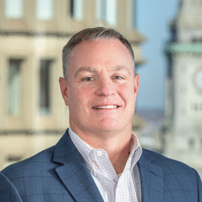 A man in a blue blazer and checked shirt stands smiling in front of a window with an urban cityscape and clocktower in the background.
