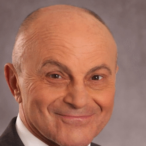 A middle-aged man with a bald head wears a dark suit, white shirt, and patterned tie, smiling slightly at the camera against a neutral gray background.