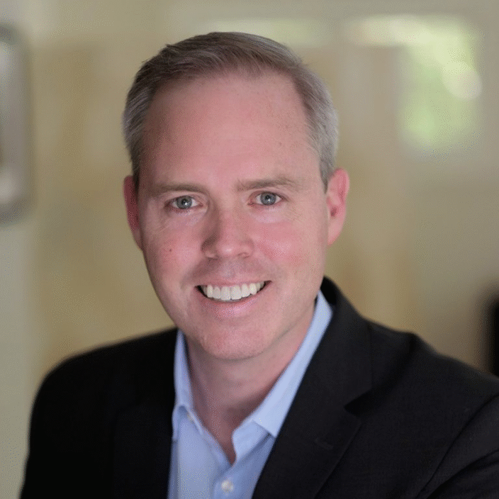 A man with short light hair wearing a dark suit jacket and light blue shirt smiles at the camera in a softly lit indoor setting.