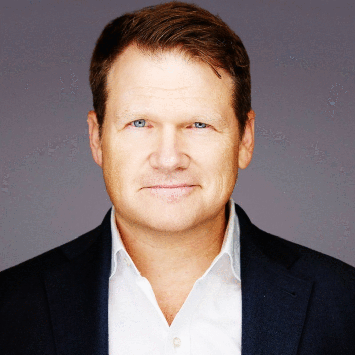 A man with light skin, short brown hair, and blue eyes is wearing a white shirt and dark blazer, smiling slightly while looking at the camera against a plain grey background.