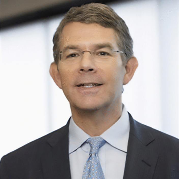 A middle-aged man with short brown hair, wearing glasses, a dark suit, a light blue shirt, and a light blue patterned tie, standing indoors in front of large windows with soft, diffused light.