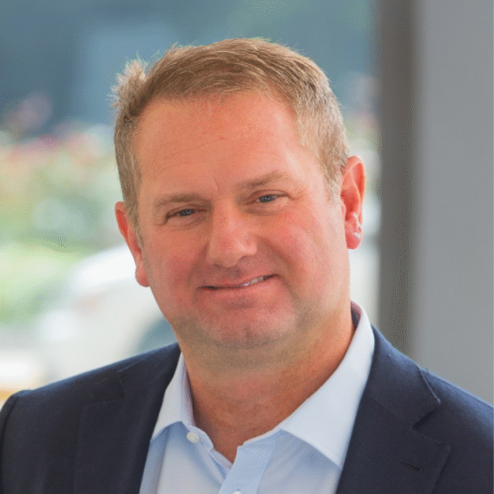 A man with short light brown hair, wearing a navy blue suit jacket and a light blue collared shirt, smiling softly while standing indoors with a blurred background.
