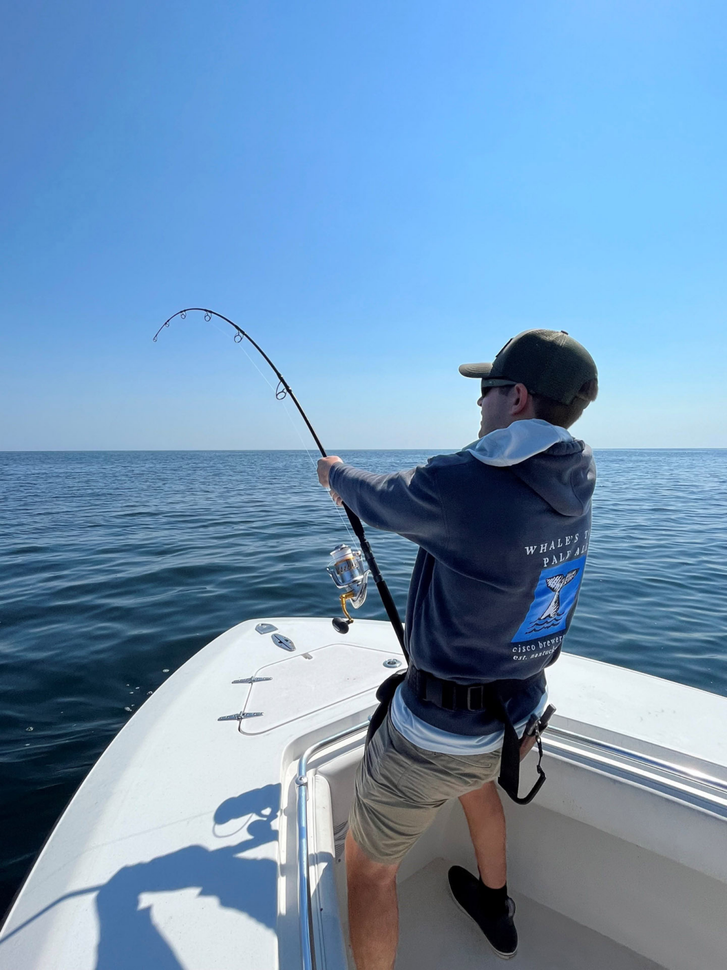 A person wearing a hoodie, shorts, and a cap is fishing on a boat in calm, open water under a clear blue sky, holding a bent fishing rod as if reeling in a catch.