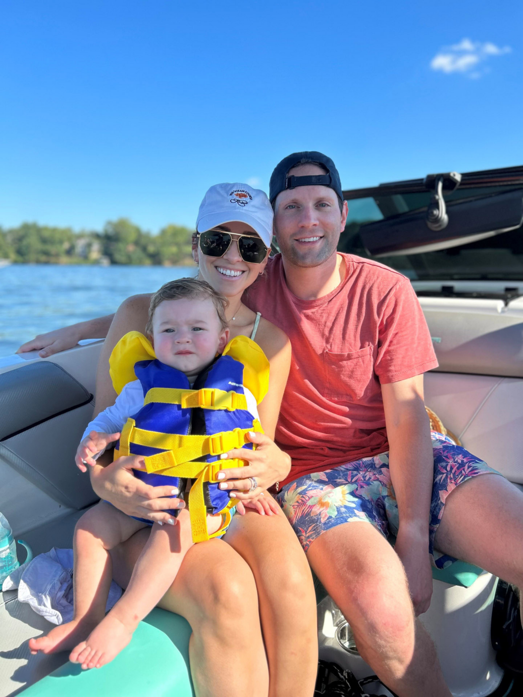 A smiling family sits together on a boat. The woman wears a white cap and yellow swimsuit, holding a toddler in a yellow life jacket on her lap. The man in a red shirt sits beside them. A lake and blue sky are in the background.