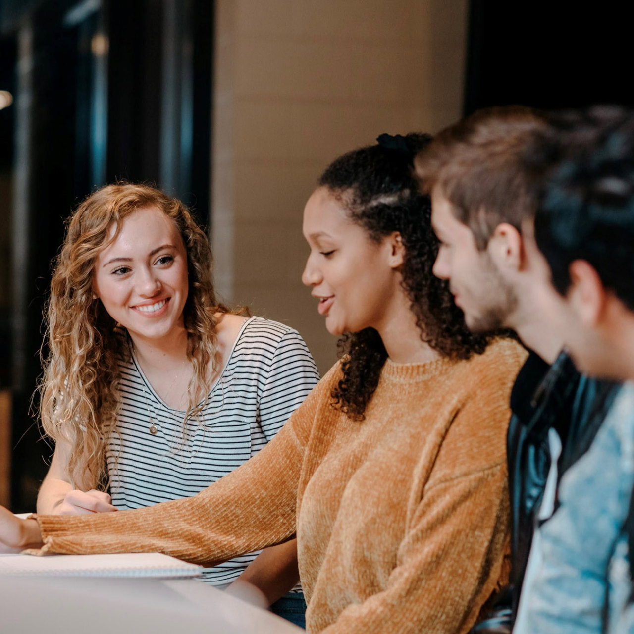 Four young adults sit at a table in a casual, modern workspace, smiling and discussing a Carnegie case study as they look at a laptop. Two women engage in conversation while two men listen attentively.