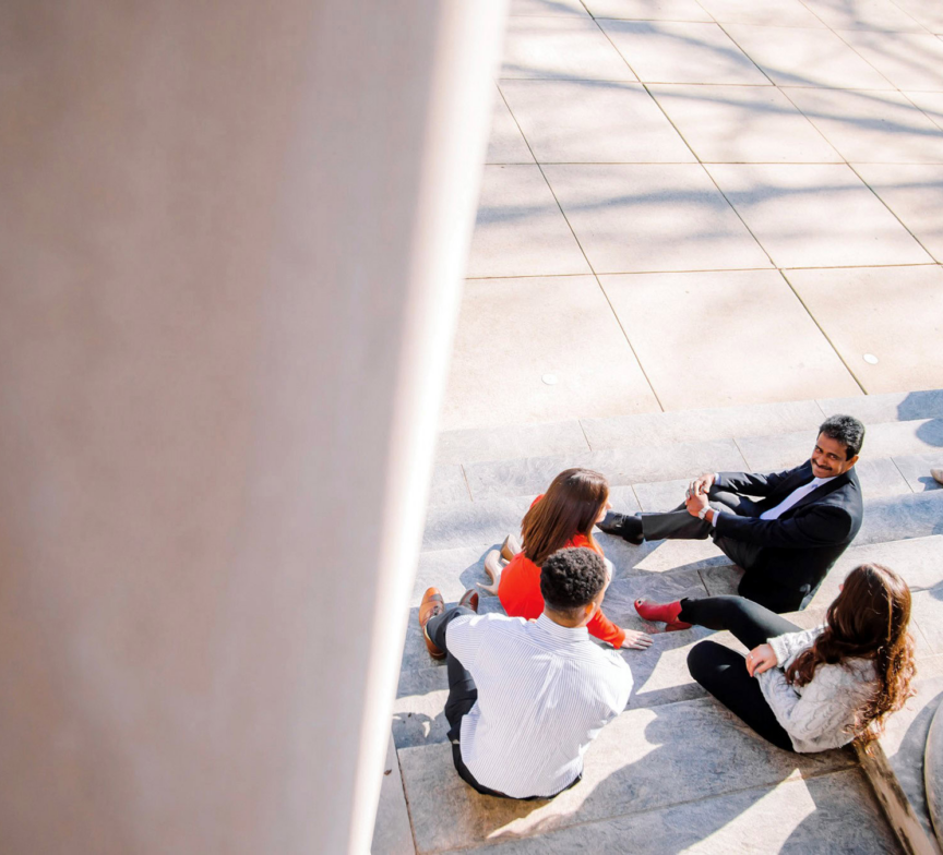 Four people sit in a small group on outdoor stone steps, engaged in conversation in sunlight—perhaps discussing a Carnegie case study—while one man wears a suit and others are casually dressed, with another person seated alone nearby.