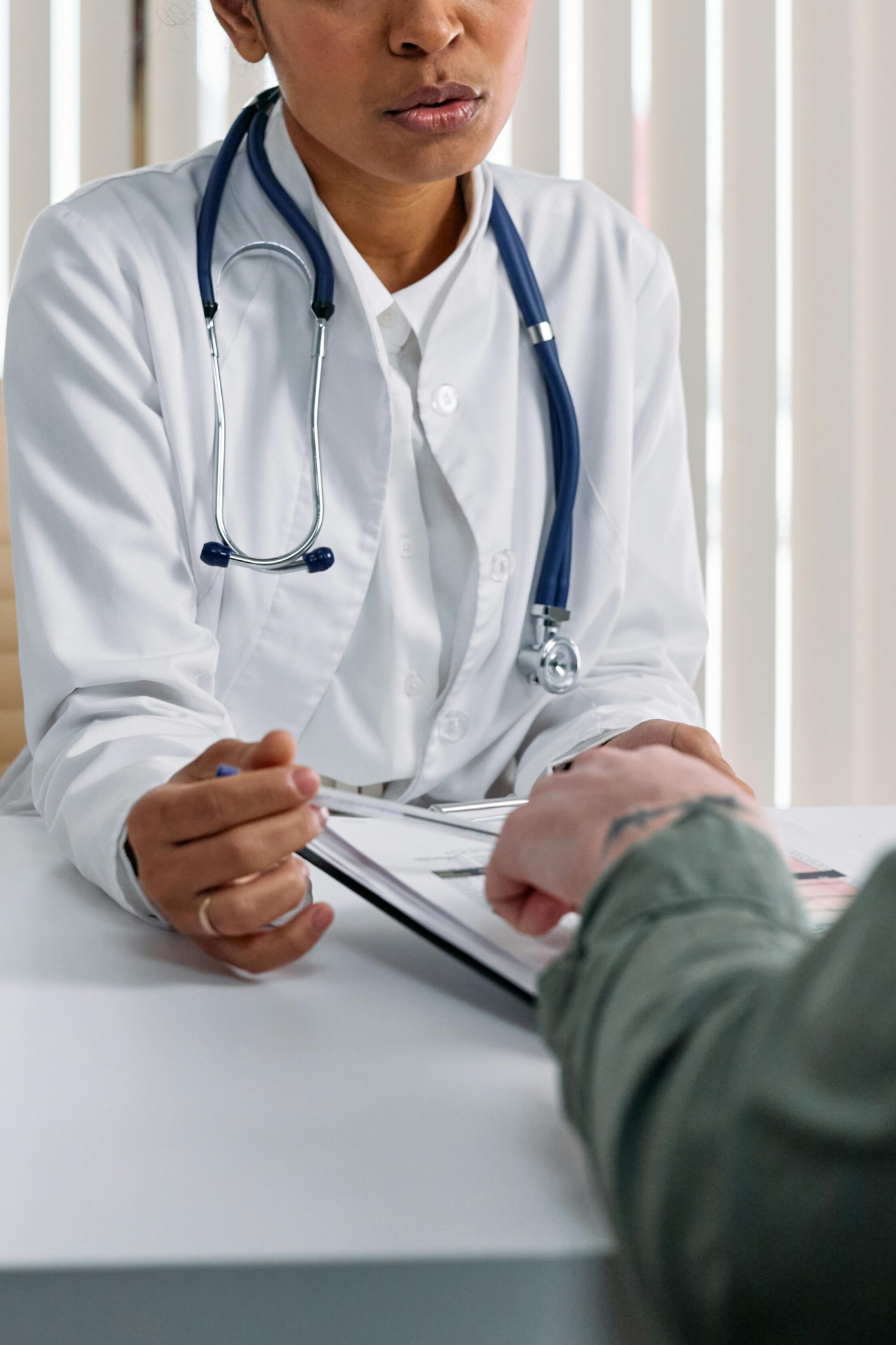 A doctor in a white coat with a stethoscope around their neck holds a clipboard and speaks with a patient across a desk in a medical office, showcasing Our Difference in compassionate care. The patient’s hand rests on the table.