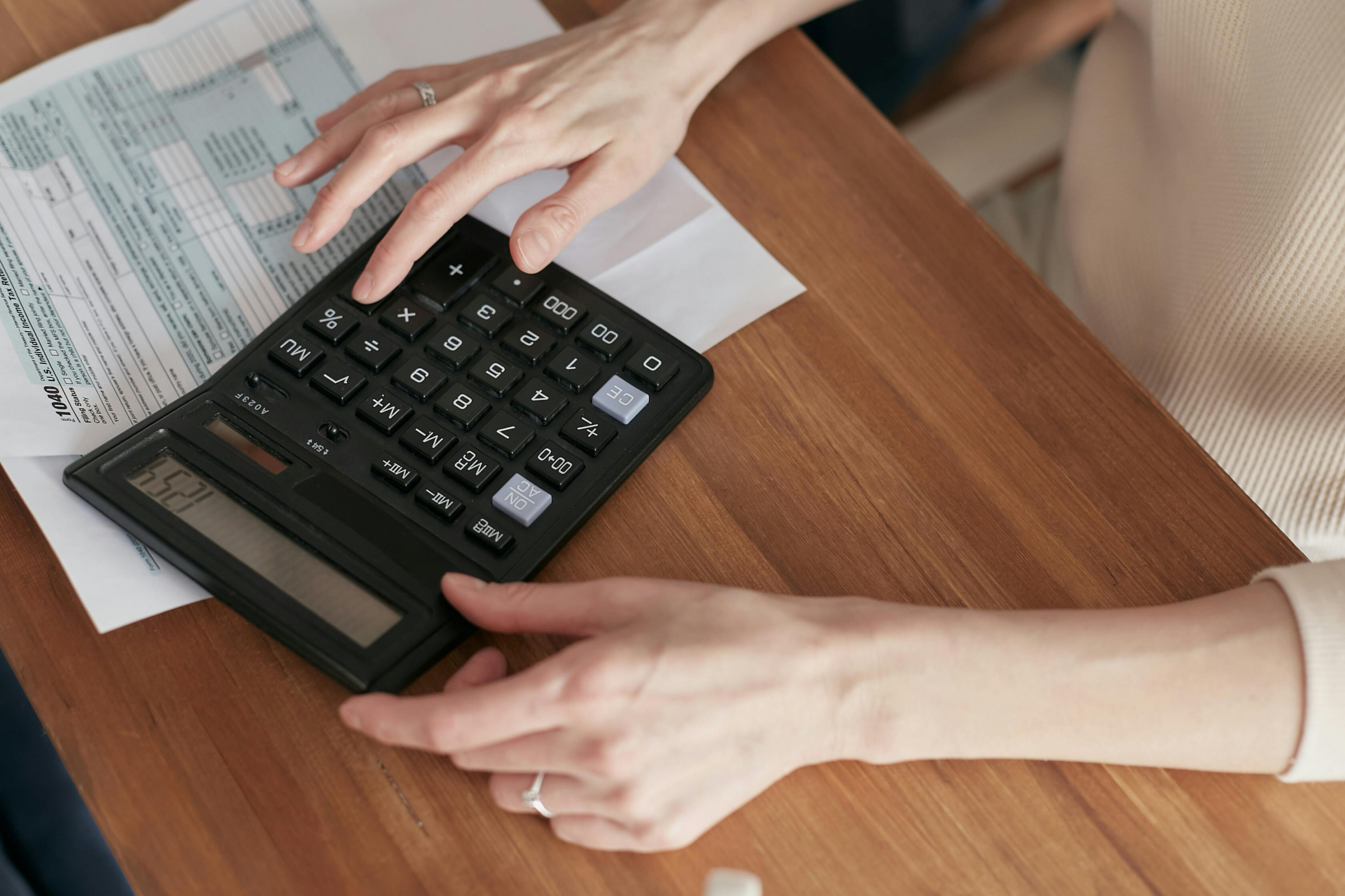 Close-up of a person using a large calculator on a wooden table with documents and paperwork underneath, focusing on their hands as they press buttons and review financial information.