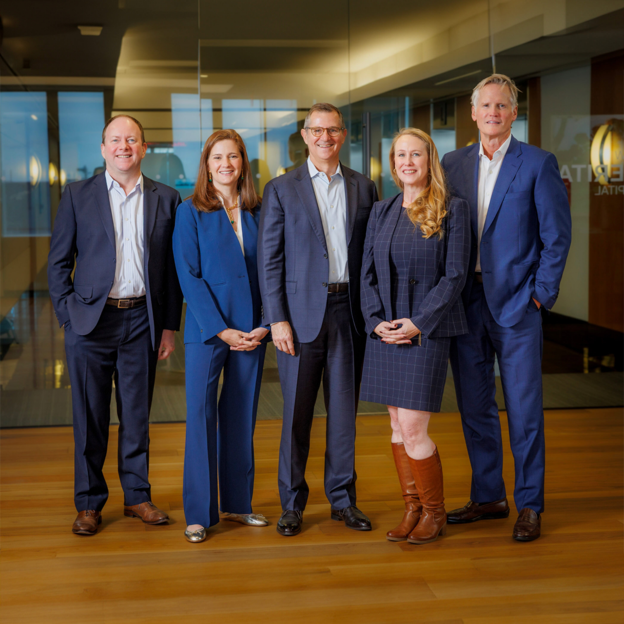 Five business professionals stand side by side in a modern office hallway, all wearing business attire, smiling and facing the camera—a welcoming scene for founders. The background features glass walls and wooden floors.