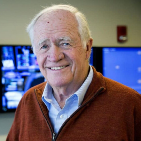 An older man with white hair smiles at the camera, wearing a rust-colored sweater over a blue collared shirt. He stands in front of large monitors displaying blue screens in an office setting.