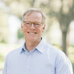 An older man with glasses and gray hair smiles outdoors, wearing a light blue button-down shirt. The background is blurred with green trees and soft sunlight.