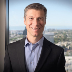 A man in a dark suit and light blue shirt smiles while standing in front of a large window with a blurred cityscape in the background.
