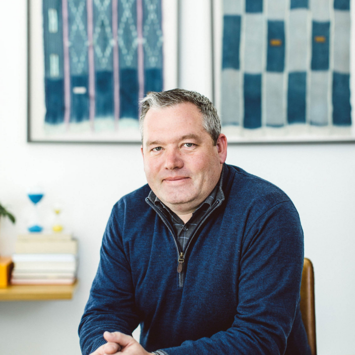 A man with short gray hair and a navy pullover sits at a wooden table, hands clasped. Behind him are two abstract blue and white framed artworks on a white wall. A stack of books and an hourglass sit on a shelf.