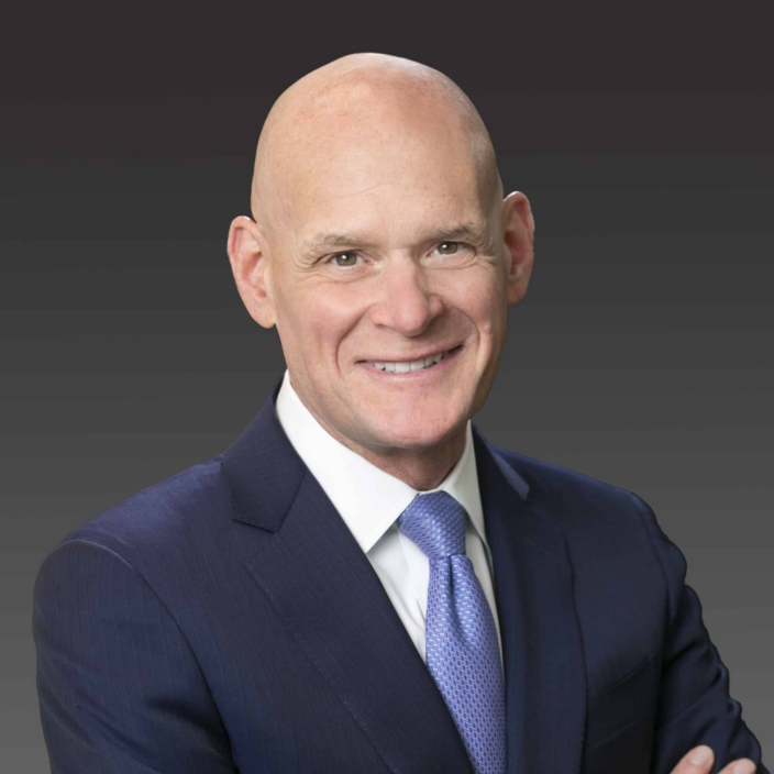 A bald man in a dark suit, white shirt, and blue tie smiles at the camera with arms crossed, set against a dark gradient background.