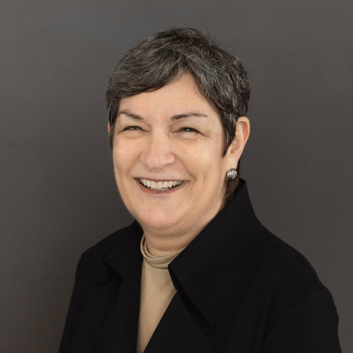 A middle-aged woman with short gray hair smiles warmly, wearing a black jacket and beige top, posing against a plain dark background.