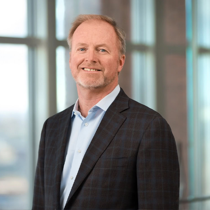 A middle-aged man with short light hair and a trimmed beard, wearing a blue dress shirt and a dark checked blazer, stands smiling in front of large windows in a modern office setting.
