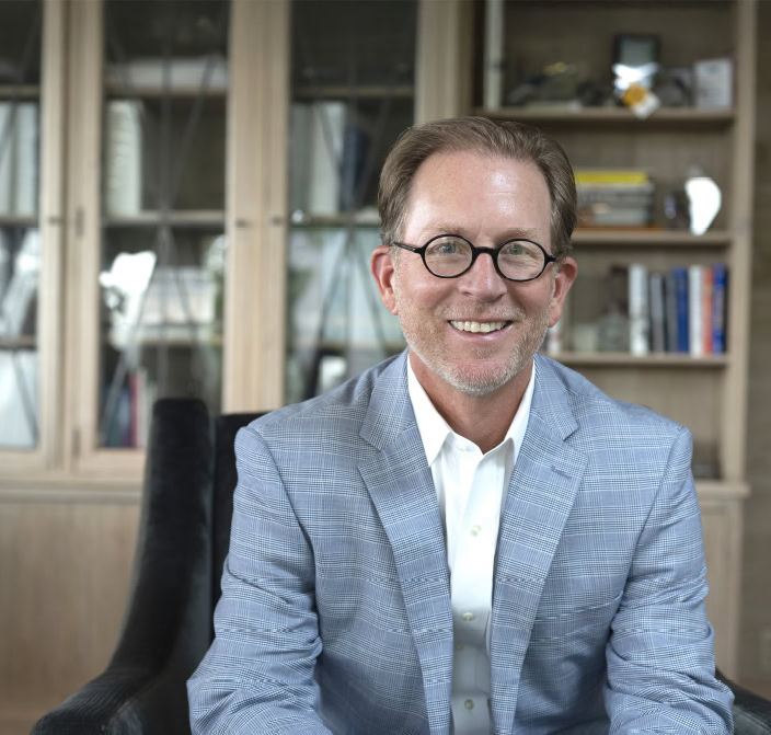A smiling man with glasses and a light blue plaid suit jacket sits in a chair in front of bookshelves with glass doors.
