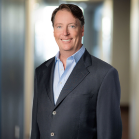 A man in a dark suit jacket and light blue shirt is smiling while standing in a blurred office hallway.