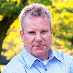 A middle-aged man with short, curly gray hair and a light blue collared shirt stands outdoors, looking directly at the camera, with a background of green trees and sunlight.