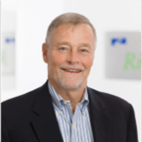 A smiling older man with short gray hair, wearing a dark suit jacket over a blue and white striped shirt, stands in an office setting with blurred signs in the background.