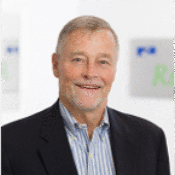 A smiling older man with short gray hair, wearing a dark suit jacket over a blue and white striped shirt, stands in an office setting with blurred signs in the background.
