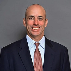 A man with a bald head smiles at the camera, wearing a dark suit jacket, light blue shirt, and patterned tie, standing in front of a plain gray background.