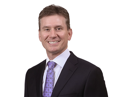 A man in a dark suit, white shirt, and patterned purple tie smiles at the camera against a plain white background.