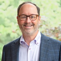 A smiling man with glasses and a short beard, wearing a checked blue blazer and a light-colored shirt, standing outdoors with a blurred green background.
